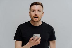 Horizontal shot of surprised startled bearded adult man holds mobile phone reacts on shocking news dressed in casual black t shirt uses modern cellular for online communication poses indoor. photo