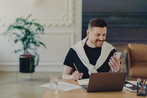 Happy bearded young man studies indoor with modern gadgets writes down notes from internet website makes records in notebook holds smartphone poses at coworking space cozy interior. Hipster student photo