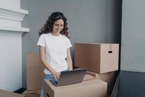 Happy european woman unloading cardboard boxes at new place and typing on laptop on the box. photo