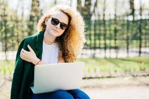 Portrait of cute young woman with curly blonde hair wearing sunglasses, white T-shirt and green jacket holding laptop on knees working at her future project isolated over green nature background photo