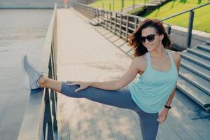 la mujer morena atlética estira las piernas usa gafas de sol camiseta polainas y zapatillas de deporte tiene una figura delgada se prepara para el trabajo al aire libre lleva un estilo de vida deportivo. flexibilidad de las personas y concepto de ejercicio foto