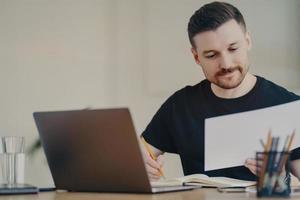 Skilled professional male expert busy doing paper report edits online files via laptop works at desktop focused at documents dressed in casual black t shirt. Bearded freelancer at home office photo