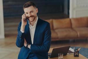 Smiling male entrepreneur talking on mobile phone while leaning on office desk photo