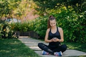 Outdoor shot of athletic woman rests after workout sits crossed legs in park surfs social networks dressed in sportswear listens music via wireless earphones uses sport app enjoys good weather photo