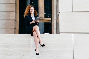 Beautiful elegant businesswoman in formal clothes and high-heeled shoes, having wavy hair and slender legs, using modern tablet for her work, looking at camera with happy look, smiling broadly photo