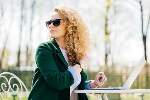 Young pretty female with fluffy blonde hair wearing sunglasses sitting on in the sun in front of laptop translating textes looking around noticing her best friend enjoying sunny weather in the yard photo