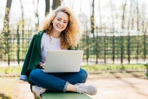 Pleased European woman having beautiful fluffy blonde hair wearing jeans, jacket and sport shoes sitting crossed legs using laptop computer for communication enjoing nature outside. People and leisure photo