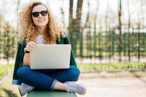 Horizontal portrait of beautiful teenager female with blonde curly hair wearing sunglasses, jeans, sport shoes sitting crossed-legs on bench in park holding laptop on her knees having happy expression photo
