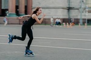 Holidays and active lifestyle concept. Slim healthy European woman rollerskates on high speed photogaphed in motion dressed in black clothing poses against blurred street background on road. photo