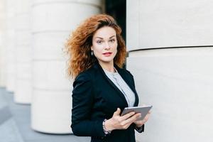 Adorable woman with curly hair wearing black suit and white blouse, holding tablet computer, typing messages or searching Internet while standing outdoor. Pretty businesswoman with modern device photo