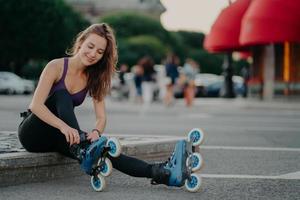 Outdoor shot of young sporty woman in sportswears puts on roller blades poses on road going to ride rollers during sunny day ties shoelaces has good mood. Recreation hobby lifestyle concept. photo