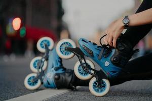 Cropped shot of unrecognizable woman puts on rollerblades poses outdoors on asphalt against blurred background. Unknown person wears rollerskates spends free time actively tries new purchase photo