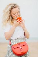Vertical portrait of lovely young woman has light curly hair, holds modern cellular, happy to see photo of lover in social networks, carries small bag, stands outdoor at coastline. Lifestyle concept