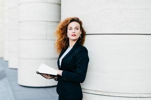 Pretty elegant woman with curly wavy hair dressed in black suit, holding pocket book in hands looking aside into distance, thinking over her future plans. Fashionable lady writing necessary notes photo