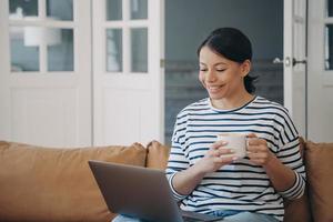 chica sonriente con una taza de café viendo una película o comunicándose por videollamada en una laptop en el sofá de casa foto