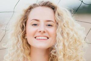 Close up shot of curly lovely young female has broad shining smile, looks positively at camera, has joyful expression, stands outdoor, has fresh skin, being glad to be photographed. Headshot photo