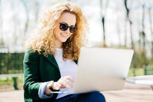 Young woman with sunglasses having fluffy hair smiling joyfully resting in park browsing internet using laptop computer surfing in social networks. People, lifestyle, technology and rest concept