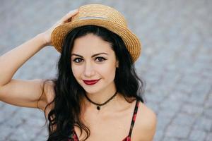 retrato al aire libre de mujer morena con ojos oscuros, labios rojos y piel sana con sombrero de verano, manteniendo la mano en la cabeza, descansando mientras camina por la calle de la ciudad antigua. juventud, concepto de moda foto