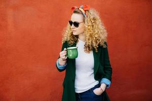 retrato de mujer bonita con cabello rizado con diadema, gafas de sol elegantes y chaqueta verde sosteniendo una taza de té verde en una mano y la otra mano en el bolsillo mirando a un lado aislado sobre una pared naranja foto