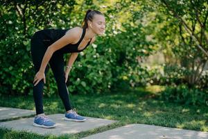 Fitness woman in black sportsclothes keeps hands on knees leans forward exercises outdoors against green nature background listens music from favorite playlist. Weight loss and workout concept photo