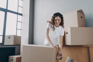 Happy girl moves alone. Young woman is packing boxes with adhesive tape for shipping. photo