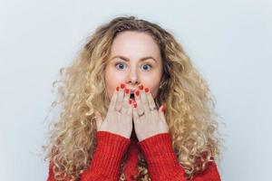Emotional lovely woman with unexpected look covers mouth with hand, has red manicure, curly hair, isolated over white studio background. Shocked young cute female surprised to recieve present photo