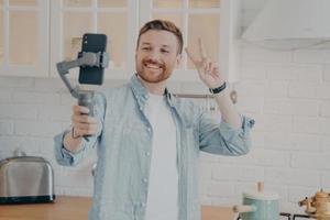 apuesto joven brunete sonriente con rastrojo haciendo selfie en la cocina foto