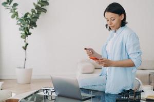 Female holds bank credit card, phone uses online banking service at workplace. E-business, ecommerce photo