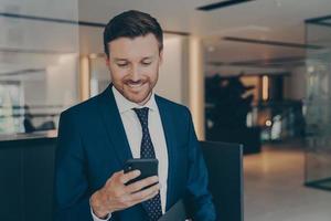 feliz joven hombre de negocios usando un teléfono inteligente parado en el pasillo de la oficina foto