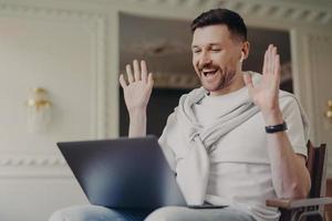 Happy male blogger talks with followers waves palms at camera of laptop computer expresses joy talks during online stream uses wireless earphones enjoys friendly conversation hears good news photo