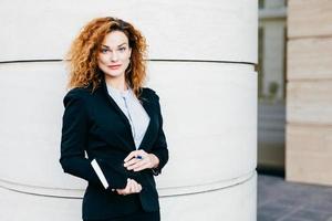 Pleasant-looking curly woman dressed formally, holding notebook in hands, going to have meeting wit her business partners. Gorgeous business lady writing in diary book. Professionalism concept photo