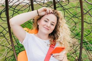 Beautiful delighted female blogger with curly light hair, uses smart phone for posting new posts on her web site, rests in hanging chair, uses wireless internet connection. People, recreation concept photo