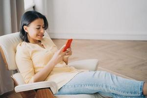 Female using phone sitting in comfortable armchair at home, shopping online. Mobile apps advertising photo