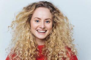 primer plano de una mujer alegre y adorable con cabello rizado, piel sana, amplia sonrisa y dientes blancos perfectos, feliz de pasar tiempo libre con los mejores amigos en compañía divertida, posa contra la pared blanca del estudio foto