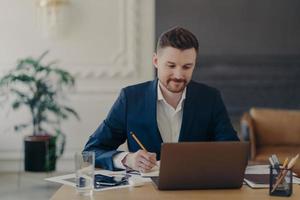 Indoor shot of handsome male office worker rewrites information from notepad in diary sits at desk dressed in formal clothes spends time for searching info. Businessman takes notes does paper work photo