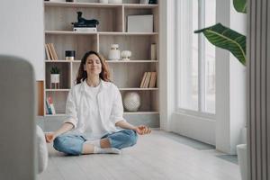 Calm happy latin woman is practicing yoga. Girl is sitting near window in morning in lotus asana. photo