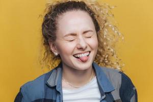 Photo of funny overjoyed female has fun alone, laughs joyfully and shows tongue, keeps eyes shut, expresses postiveness, isolated over yellow background. People and facial expressions concept