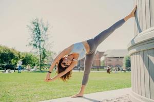 mujer delgada y flexible hace ejercicios de estiramiento al aire libre se mantiene en buena forma física vestida con top recortado y polainas se para en una pierna se inclina con los brazos levantados se calienta antes del entrenamiento cardiovascular foto