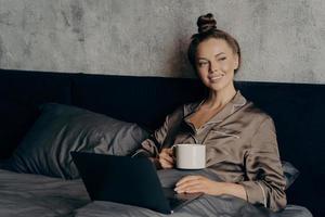 Smiling beautiful young female using laptop while lying in bed with cup of coffee photo