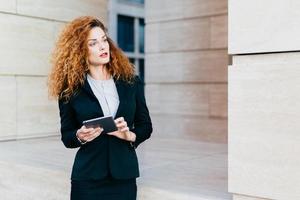 Female entrepreneur with curly bushy hair wearing formal clothes, holding modern tablet, looking with pensive expression aside, planning her working day. Woman developing project using touch pad photo