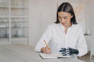 Disabled european girl with cyber arm is taking notes at the desk and doing homework. photo