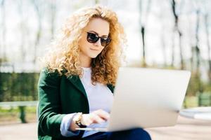 retrato de una linda mujer con cabello rubio esponjoso y piel pura con gafas de sol y chaqueta verde concentrada en la pantalla de su laptop leyendo atentamente historias interesantes sentadas en el banco foto