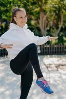 Fitness sport healthy lifestyle concept. Pleased sporty woman tries to balance on one leg keeps knee raised has interval training or cardio workout wears white sweatshirt leggings and sneakers photo