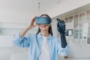 Disabled person gets rehabilitation. Handicapped girl with cyber arm in vr goggles at home. photo