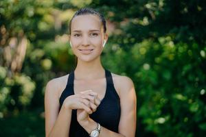 Outdoor shot of pretty dark haired woman keeps hands together being in good physical shape as goes in for sport regularly wears sportsclothes listens music while having workout at green park photo