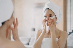 Girl applies eye patches and looking into mirror. Attractive woman wrapped in towel after bathing. photo