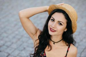 Portrait of lovely young female with dark luxurious hair, appealing eyes, red lips smiling gently while posing at camera wearing straw summer hat and dress keeping her hand on head resting outdoor photo