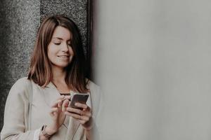 chica de aspecto agradable con corte de pelo a la moda, pasea afuera, envía mensajes por teléfono celular, disfruta de la conexión a Internet en roaming, usa una elegante chaqueta blanca, usa la aplicación, chatea con amigos foto