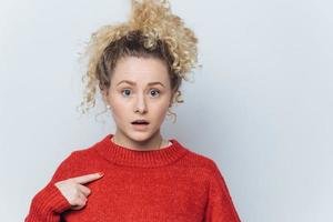 Shocked stunned young woman with surprised expression, indicates at blank red sweater, advertizes new outfit, poses against white studio background with copy space for your promotional text. photo