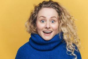 Stunned beautiful blue eyed young woman with light crisp hair, doesnt expect to recieve shocking news, wears knitted sweater, stares at camera with bugged eyes, isolated over yellow background photo